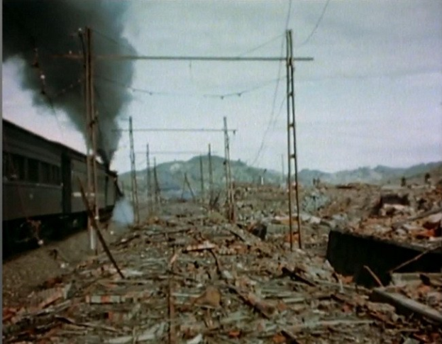 Ein Zug fährt durch ein beschädigt Gebiet mit Rauch, der aus ihm aufsteigt, umgeben von Masten und Dröhten, mit Höhen und einem klaren Himmel im Hintergrund und verstreuten Trömmern am Boden.