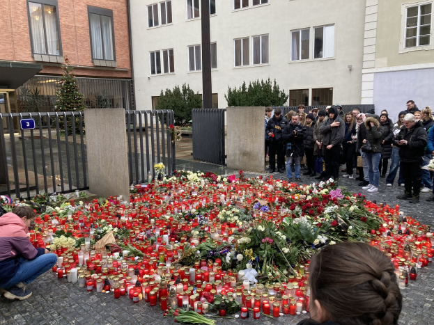Eine Gruppe von Menschen an einer improvisierten Gedenkstätte mit Blumen, Kerzen und Gegenständen auf dem Boden, einschließlich einer Person, die kniet, während sie eine Kamera hält; Gebäude, Pflanzen, ein Pfahl und ein Tor sind im Hintergrund sichtbar.