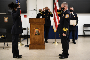 Eine Gruppe uniformierter Personen in Masken steht vor einem Rednerpult mit Mikrofon, umgeben von Fahnen, Stühlen und Tischen, während offenbar eine Vereidigung eines neuen Feuerwehrchefs stattfindet.
