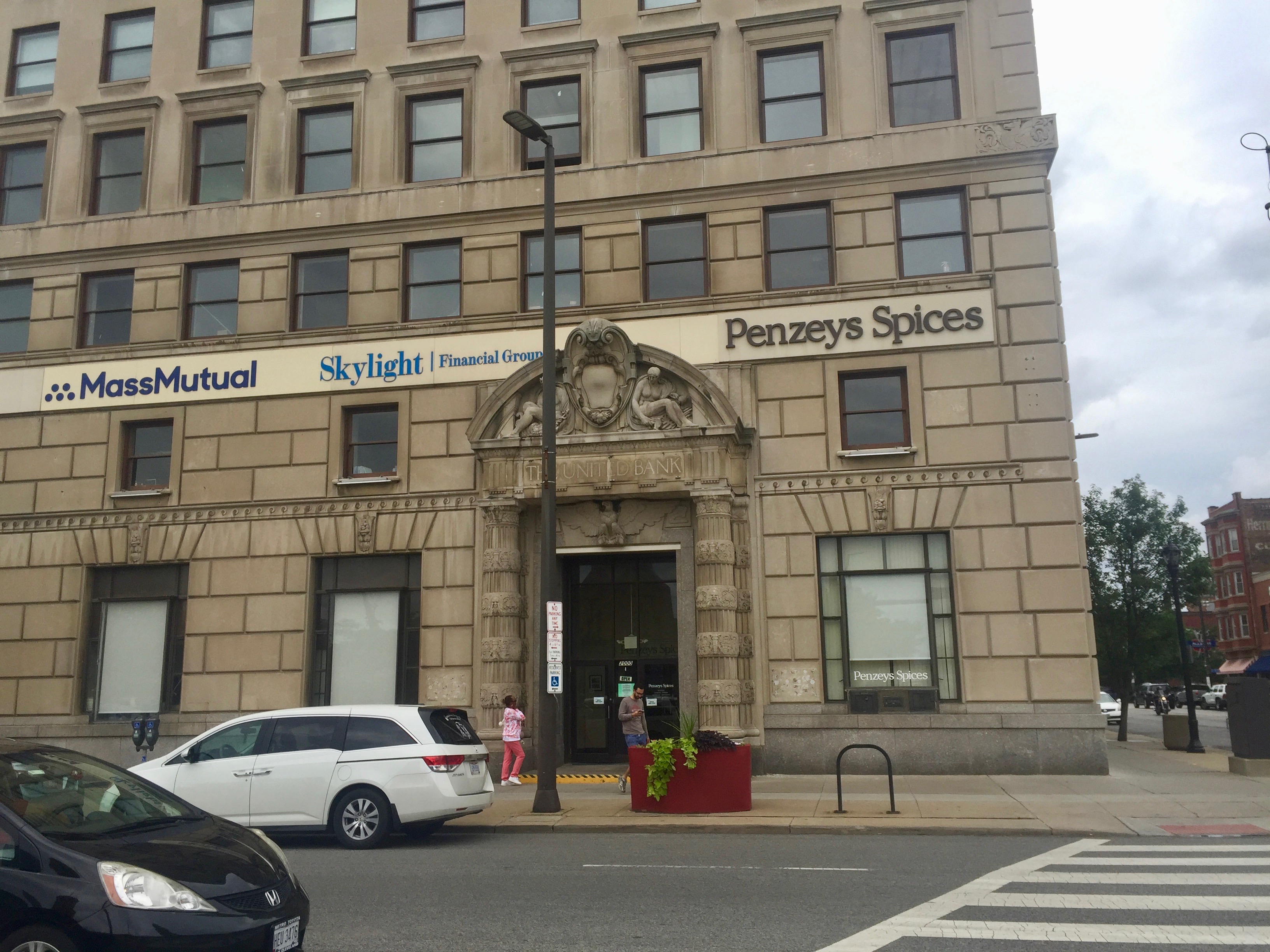 Ein großes Gebäude, das als Hauptsitz der Massachusetts Mutual Financial Group identifiziert wurde, mit parkenden Autos davor, Straßenlaternen, Strommasten, Kraftfahrzeugen auf der Straße, Personen auf dem Gehweg, Topfpflanzen, Bäumen und einem bewölkten Himmel im Hintergrund.