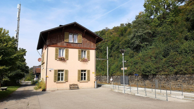 Ferienhaus zum Verkauf in Baden-Württemberg, Deutschland, mit einem mehrfenstrigen Gebäude mit einer vorderen Einfriedung, umgeben von Straßeninfrastruktur und Grün unter einem bewölkten Himmel.