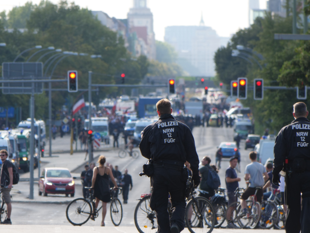 Zwei Polizisten stehen neben einer Gruppe von Radfahrern auf der Straße, Autos, Verkehrszeichen, Bäume, Gebäude und ein klarer blauer Himmel im Hintergrund.