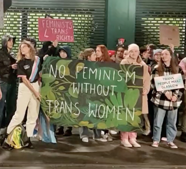 Eine Gruppe von Menschen hält ein Transparent mit der Aufschrift "Kein Feminismus ohne Transfrauen" und verschiedene Schilder in der Öffentlichkeit.