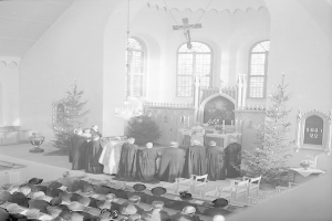 Eine Gruppe von Menschen in Schwarz-Weiß-Kleidung sitzt auf Stühlen vor einem Chor in einer Kirche, mit geschmückten Weihnachtsbäumen, Kerzenständern, einem Kreuz an der Wand und Fenstern im Hintergrund.