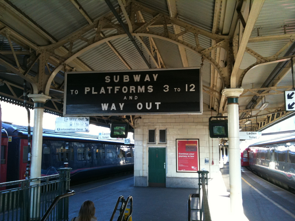 Zwei Züge neben einem Bahnsteig mit zwei Bildschirmen, einer Person in der Nähe einer Treppe, einer Tür an der Wand und mehreren Schildern an der Decke.