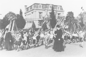 Schwarz-weißes Bild einer Gruppe von Menschen, die eine Straße entlanggehen und Fahnen halten, mit Hüten, einem Maibaum, Bäumen, Gebäuden und Himmel im Hintergrund.
