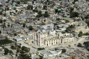 Luftaufnahme einer Stadt mit einer beschädigten Kirche im Zentrum, umgeben von Gebäuden, Bäumen, Straßen, Fahrzeugen und Erdbewegungen.