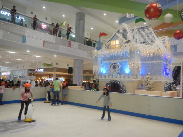 Eine Gruppe von Menschen, die auf einer Eisbahn in einem Einkaufszentrum Schlittschuh laufen, umgeben von einem häuslichen Aufbau mit Lichtern und festlichen Dekorationen, wobei Zuschauer und zusätzliche Dekorationen im Hintergrund zu sehen sind.