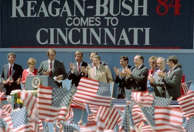 Gruppe von Menschen auf einer Bühne, die klatschen, mit einem Podium, Mikrofon und Flaggen im Hintergrund, Wand mit Text.