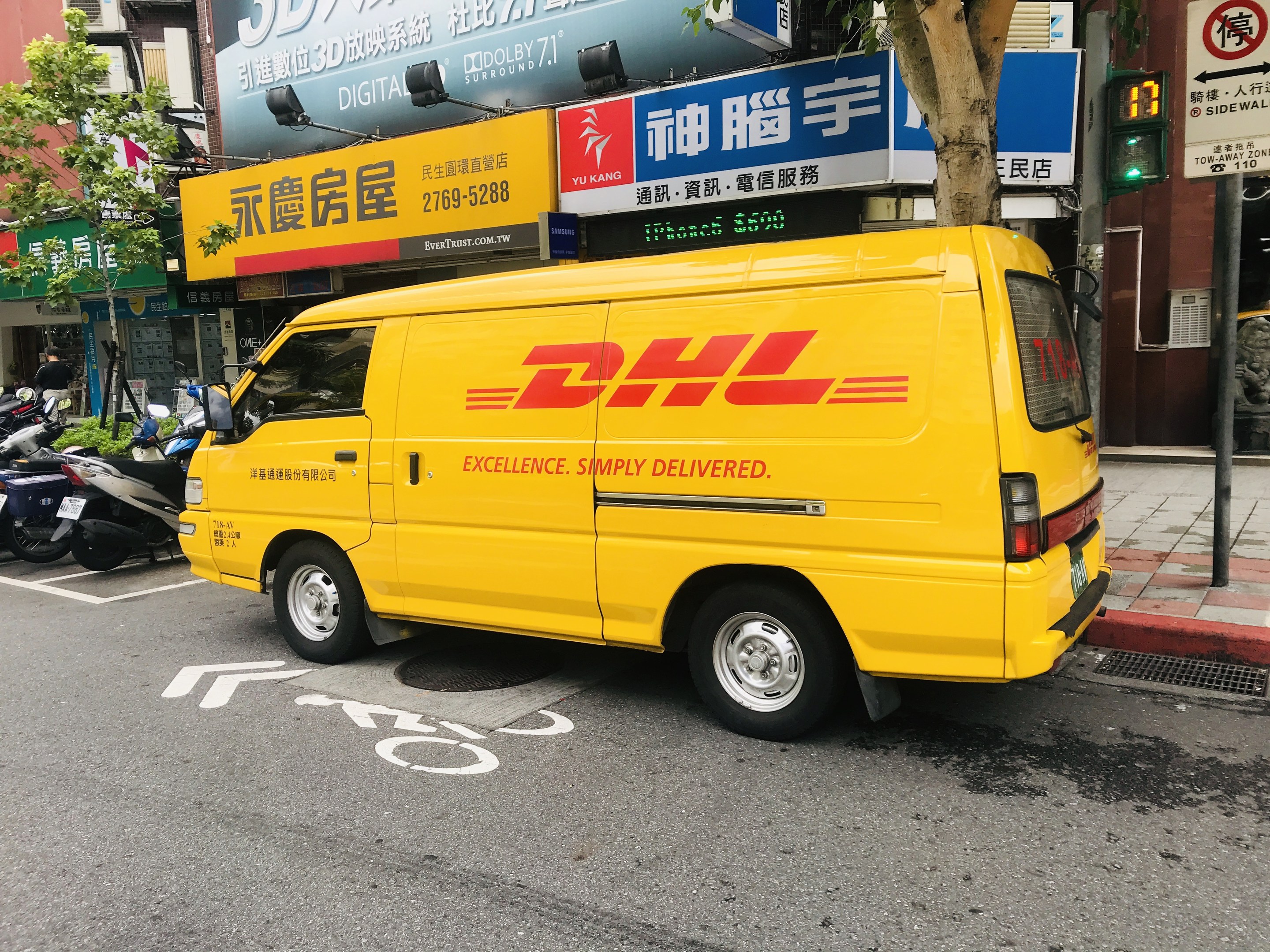 Gelbes DHL-Lieferfahrzeug auf der Straßenseite geparkt, umgeben von Gebäuden, Bäumen, Schildern, Mäständen, Ampeln und einem Fussgänger auf dem Gehweg.