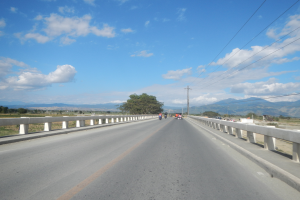 Eine Autobahn mit Motorradfahrern darauf, umgeben von Geländern, Strommästen mit Drähten, Bäumen, Hügeln und einem bewölkten Himmel im Hintergrund.