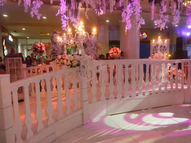 Weiße Reling mit Blumen und Kronleuchtern an der Decke bei einer Hochzeitsfeier, umgeben von Menschen, Säulen und Lichtern im Hintergrund und einem Bildschirm auf der rechten Seite.