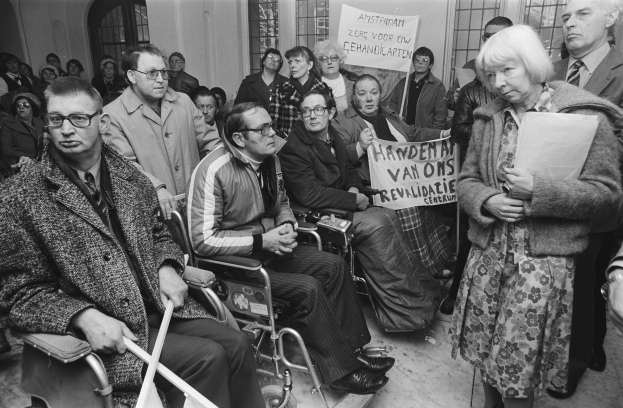 Eine Gruppe von Menschen in Rollstühlen und Stehenden vor einer Menge, mit Schildern, Fenstern und einer Wand im Hintergrund, in Schwarz-Weiß dargestellt.