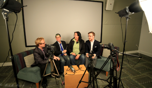 Eine Gruppe von drei Personen sitzt auf einem Sofa und gibt ein Interview, wobei eine Person links in einem schwarzen Mantel und Hose sitzt, eine Person in der Mitte einen blauen Schlips trägt und ein Mikrofon hält, und eine Person rechts lächelt. Ein Mikrofonrecorder befindet sich auf dem Tisch in der Mitte, Kameras sind auf der rechten und linken Seite und große Scheinwerfer sind vorhanden. Ein Projektionsschirm ist im Hintergrund sichtbar.