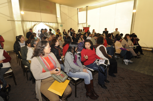 Ein voller Saal mit vielen sitzenden Personen und zwei sichtbaren Ausgängen im Hintergrund, durch die einige Menschen eintreten.