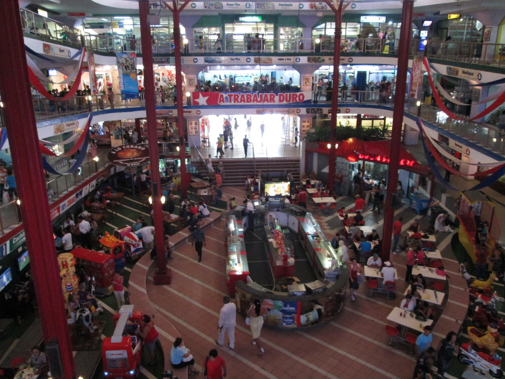 Eine belebte Einkaufszentrumszene mit Säulen, Menschen, Tischen, Fliesen, Flaggen, Geschäften, einem Banner, Treppen und einem Namensschild.