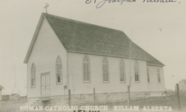 Ein Schwarz-Weiß-Foto von der St. Joseph's Lutheran Church in Killam, Alberta, das ein Haus mit einem Dach, Fenstern, Türen und einem Kreuz auf dem Dach zeigt, umgeben von einem Zaun, Gras und anderen Häusern im Hintergrund, mit Text oben und unten auf dem Bild.