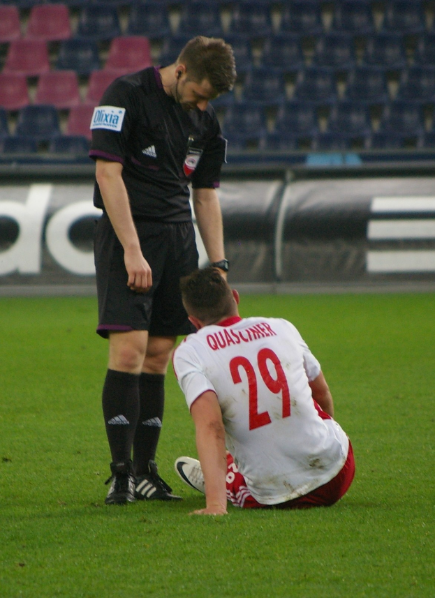 Ein Fussballer und Schiedsrichter sitzen auf dem Boden in einem Stadion, beide in Sportkleidung.