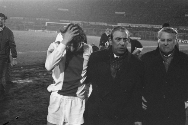 Eine Gruppe von M├Ąnnern in Not steht zusammen auf einem Fussballfeld mit einem Stadion im Hintergrund, aufgenommen in Schwarz-Wei├č.