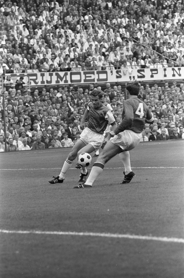 Schwarzes und weißes Foto von zwei Fußballspielern im Einsatz auf einem Feld mit Zuschauern und einem Banner mit der Aufschrift "Bobby Charlton im Einsatz für Manchester United" im Hintergrund.