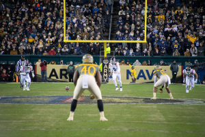 Ein Fussballspiel wird in einem Stadion mit Spielern in Sportkleidung und Helmen im Vordergrund und einer Zuschauermenge im Hintergrund gespielt, sowie sichtbaren Pfählen und Schildern mit Text.