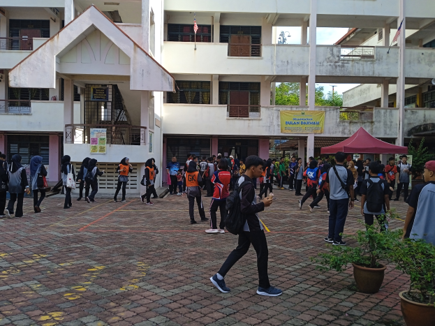 Eine Gruppe von Menschen, einige mit Taschen, geht vor einem Geb├Ąude mit Fenstern, Gel├Ąndern, Pfeilern, Bannern, Flaggen, einem Zelt, Topfpflanzen und B├Ąumen unter einem bew├Âlktem Himmel.