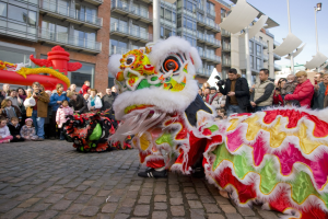 Ein lebendiges chinesisches Neujahrsfest in Amsterdam mit einem Löwen tanzen im Vordergrund und einer Menschenmenge, einige halten Kameras, vor einem Hintergrund aus Gebäuden, Laternenmasten und einem klaren blauen Himmel.