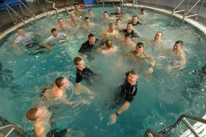 Gruppe von Männern in einem Schwimmbecken mit Armen um die Taille, umgeben von Metallstäben und einem gefliesten Boden, mit verstreuten Gegenständen im Hintergrund.