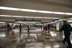 Eine Gruppe von Menschen, die durch eine belebte U-Bahn-Station gehen, mit einem Polizeibeamten im Vordergrund, Anzeigetafeln an der Decke und verschiedene strukturelle Elemente wie Geländer.