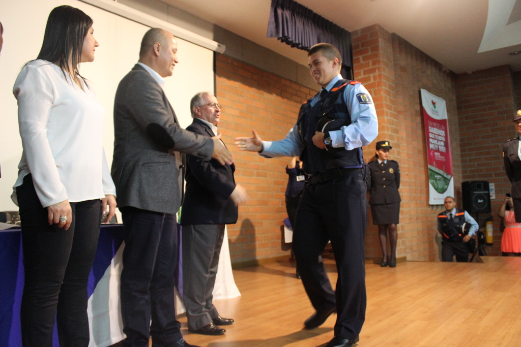 Eine Gruppe von Menschen steht auf einer B√ľhne mit einem Banner, Lautsprecher, Tisch, Bildschirm und Vorh√§ngen im Hintergrund; eine Person in Uniform h√∂lt eine M√ľtze, und eine Frau tr√§gt ein wei√ües Hemd.