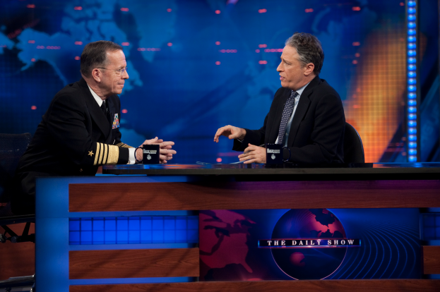 Zwei Männer in schwarzen Blazern, Hemden und Krawatten sitzen an einem Tisch mit Mikrofonen zwischen ihnen und führen ein Gespräch vor einem Fernseher und einem blauen Hintergrund.