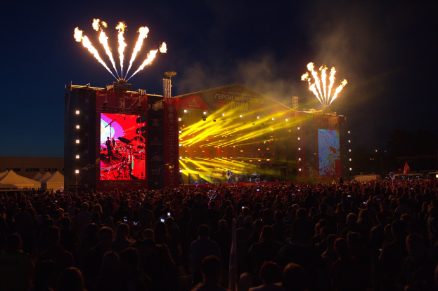 Eine große Menschenmenge bei einem Musikfestival schaut auf einer hell erleuchteten Bühne Performern zu, mit Zelten links daneben und Feuerwerk am dunklen Himmel dahinter.