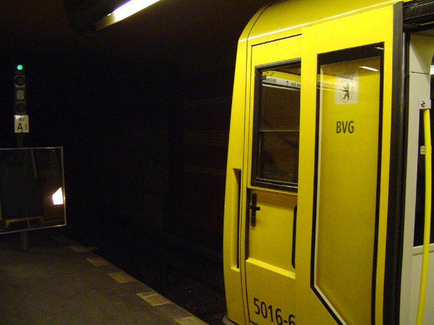 Ein Bahnhof mit einem Zug auf der rechten Seite, einem Pfahl und einer Tafel auf dem linken Bahnsteig, einer Lampe oben und einer Bahnstrecke unten.