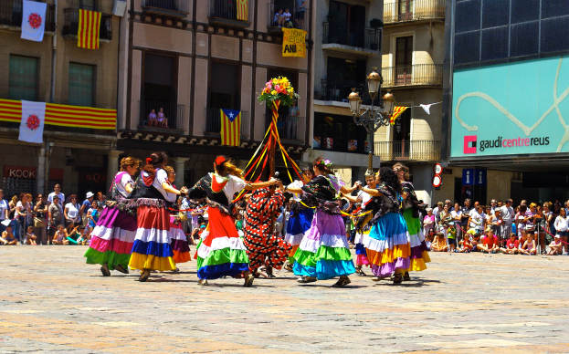 Eine Gruppe Frauen in bunten Kleidern tanzt auf der Straße, umgeben von einer Menge Menschen, mit Gebäuden, Geländern, Balkonen, Fahnen, einem Laternenmast und einer Tafel mit Schrift im Hintergrund.