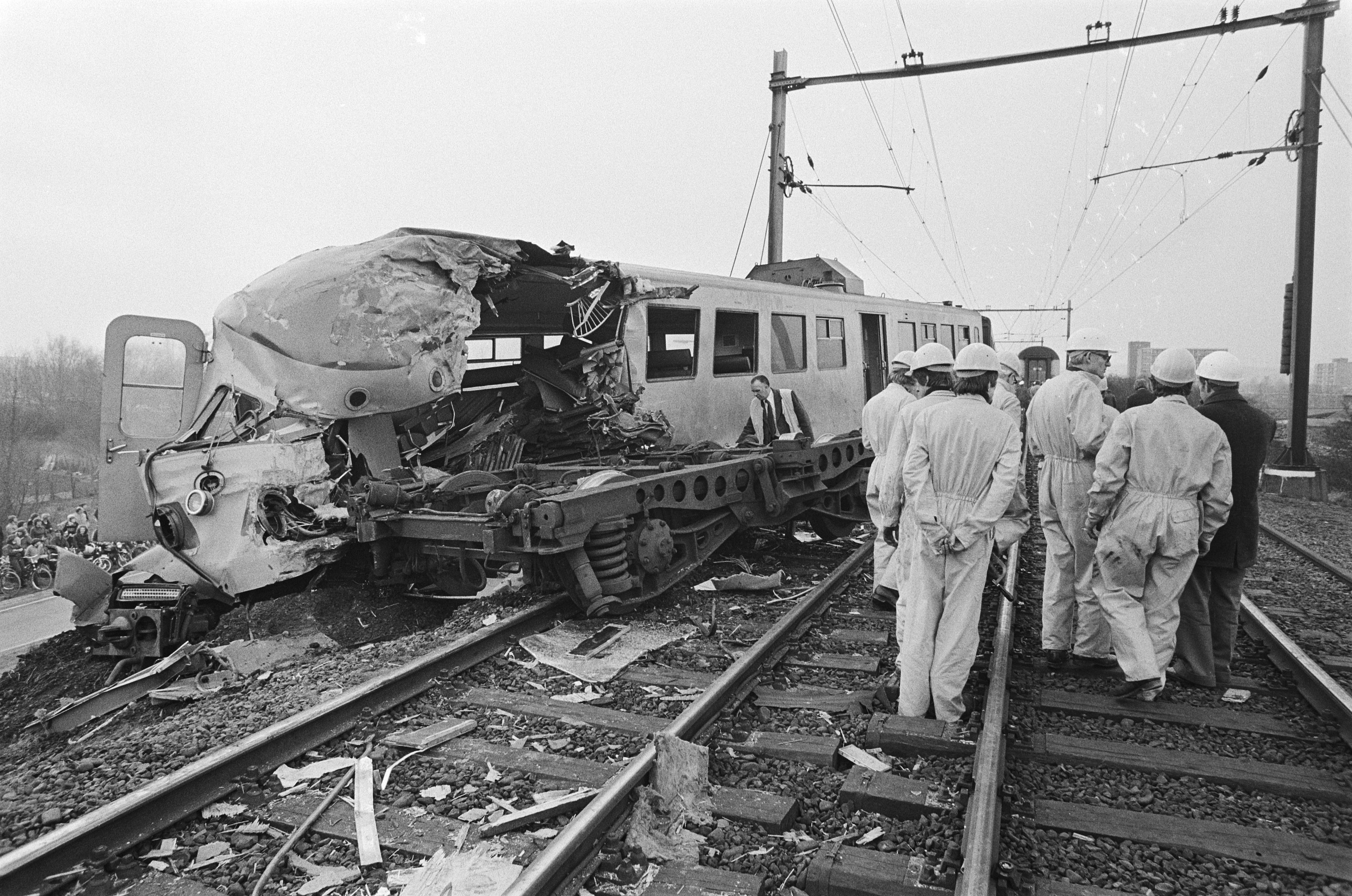 Ein Schwarz-Weiß-Bild eines Zugunglücks mit Einsatzkräften in Helmen, die um das Wrack herumstehen, vor einer Kulisse aus Strommasten, Bäumen, Gebäuden und einem klaren Himmel.