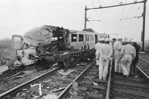 Ein Schwarz-Weiß-Bild eines Zugunglücks mit Einsatzkräften in Helmen, die um das Wrack herumstehen, vor einer Kulisse aus Strommasten, Bäumen, Gebäuden und einem klaren Himmel.