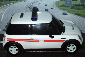 Weißes Polizeiauto mit "Polizei"-Aufschrift auf einer Landstraße, umgeben von fahrendem Verkehr, Gras und Bäumen auf beiden Seiten.