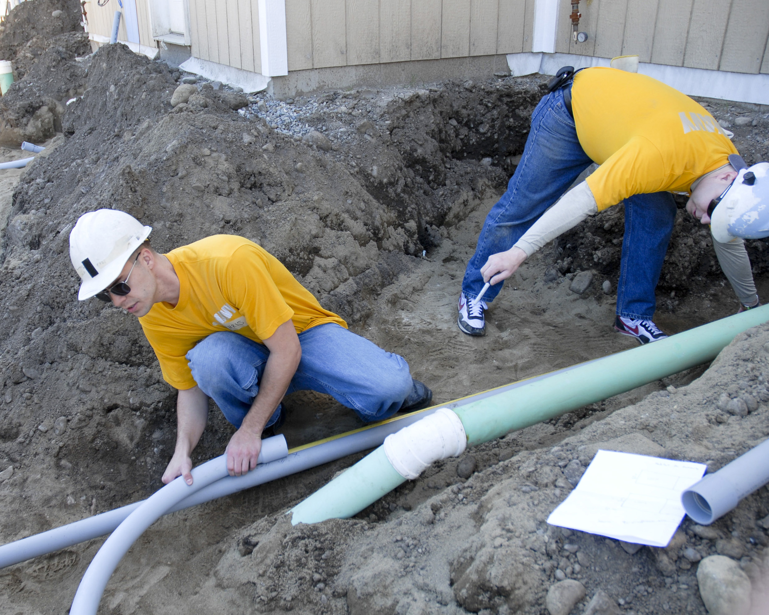 Zwei Männer in gelben Hemden und Helmen arbeiten an einem Rohr, umgeben von Erde, Steinen und anderen Gegenständen, mit einem Gebäude im Hintergrund.