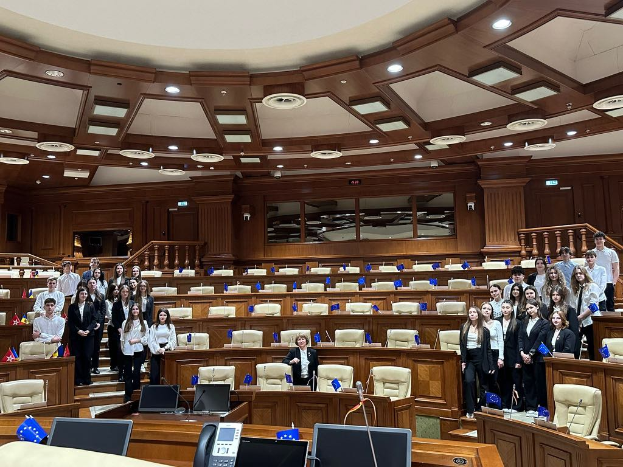 Eine Gruppe von Europaabgeordneten steht in einem großen Raum mit Tischen, Stühlen, Laptops, Mikrofonen und Flaggen, sowie Geländern und Deckenleuchten im Hintergrund.