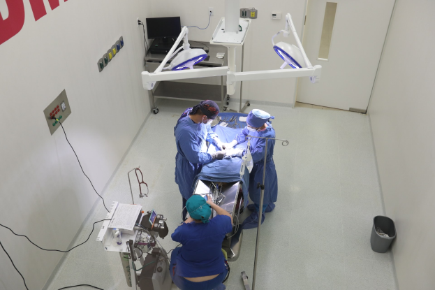 Eine Gruppe von Chirurgen führt einen Eingriff in einem Operationssaal durch, mit einem Patienten auf einer Trage umgeben von medizinischer Ausrüstung, einer beschrifteten Wand auf der linken Seite, einem Objekt auf dem Boden auf der rechten Seite und verschiedenen medizinischen Geräten im Hintergrund.