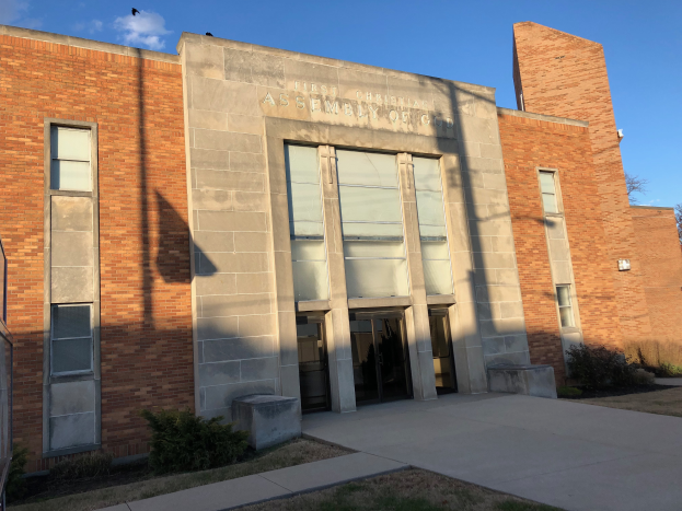 Ein Ziegelgebäude mit Glasfenstern und -türen, einem Metallzaun, Pflanzen, Gras, einem Weg, einer Baumgruppe und einem bewölkten Himmel; auf dem Gebäude steht ein Schild "Erste christliche Versammlung Gottes."
