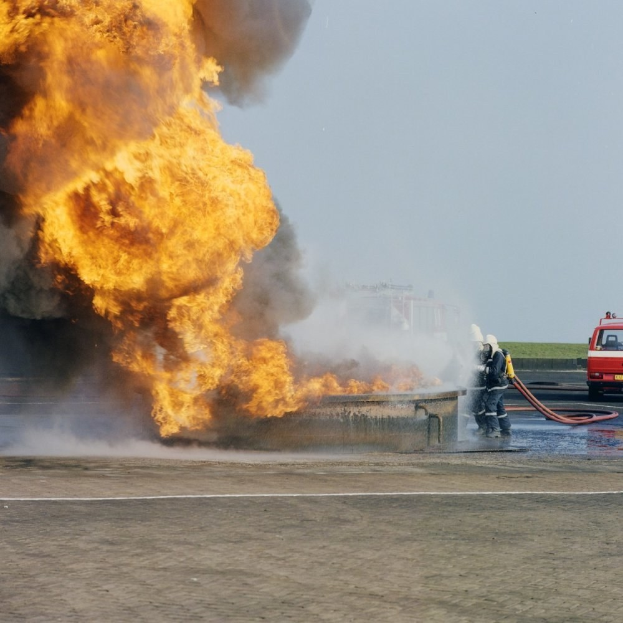 Feuerwehrlöschfahrzeug in Flammen an der Stra√enseite mit zwei Helmträgern, die Stäbe halten, einem Fahrzeug im Hintergrund und dem Himmel.