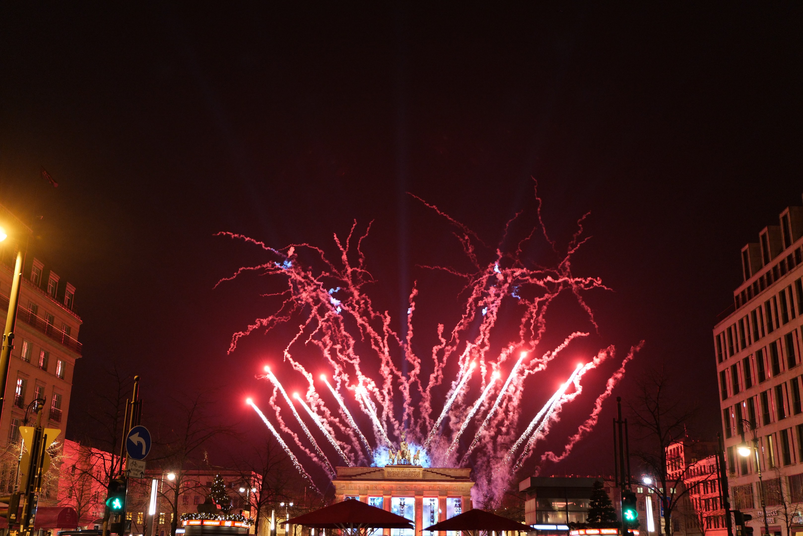 Eine belebte Stadtstraße während einer Silvesterfeier in Berlin mit Menschen, Fahrzeugen, Gebäuden und Feuerwerk, das den Nachthimmel erhellt.