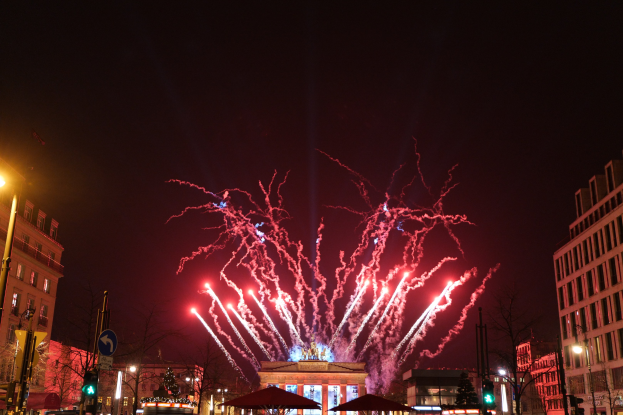 Eine belebte Stadtstraße während einer Silvesterfeier in Berlin mit Menschen, Fahrzeugen, Gebäuden und Feuerwerk, das den Nachthimmel erhellt.