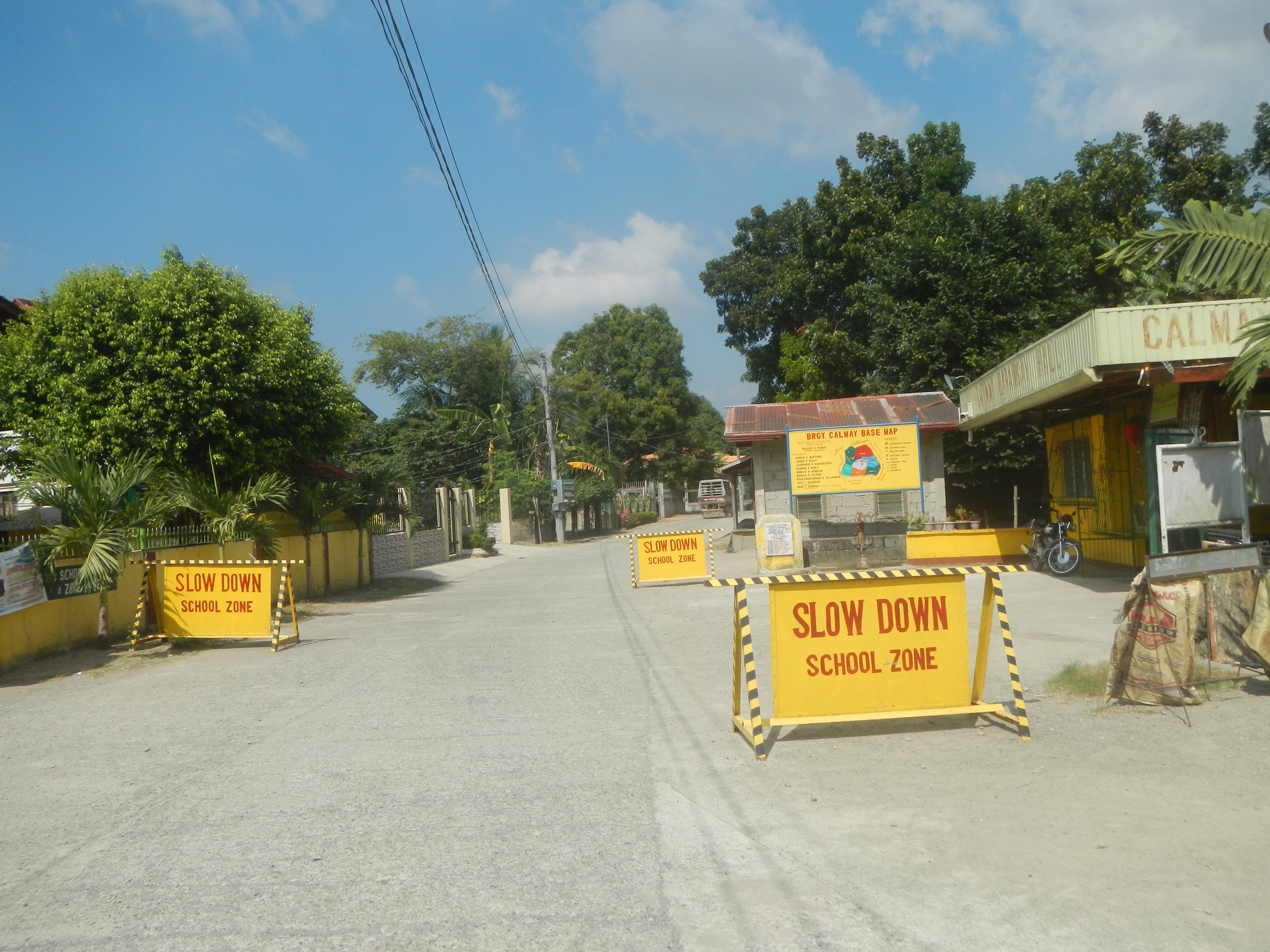 Eine Straße mit einem gelben "Slow Down School Zone"-Schild, parkenden Fahrzeugen auf der rechten Seite und Bäumen, Gebäuden, Polen, Drähten und Himmel im Hintergrund.