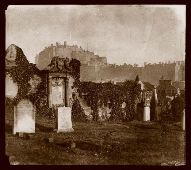 Alte Schwarz-Weiß-Fotografie eines Friedhofs mit Grabsteinen, Efeu und Gebäuden im Vordergrund und einer Burg im Hintergrund unter einem sichtbaren Himmel.