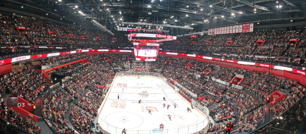Eine große Indoor-Hockey-Arena voller Zuschauer unter hellen Deckenlampen, mit einem zentralen Scoreboard und einer enthusiastischen Menge, die das Spiel anfeuert.