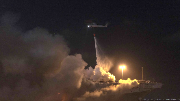 Ein Hubschrauber fliegt über ein großes Schiff mit qualmendem Rauch, beleuchtet von den Lichtern des Schiffs und dem Himmel im Hintergrund.