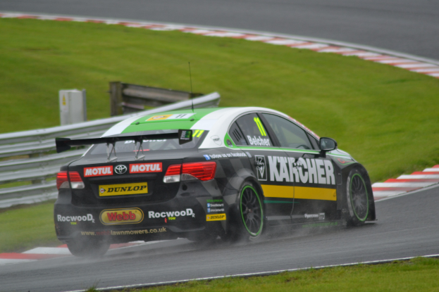 Ein Auto mit Text auf der Seite fährt auf einer regennassen Rennstrecke, die von Gras und einer Begrenzung mit Holzkonstruktion gesäumt ist.