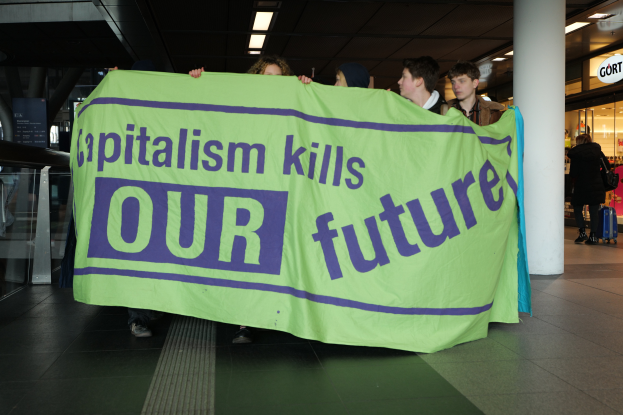 Eine Gruppe von Menschen in einem Flughafen, die ein Banner halten, auf dem "Kapitalismus tötet unsere Zukunft" steht, mit einer Säule in der Mitte und einer Glaswand links und Geschäften mit Namensschildern und Deckenleuchten im Hintergrund.
